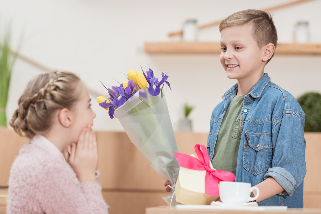 smiling boy presenting bouquet and box with ribbon to surprised girlfriend at cafe   - Photo, Image