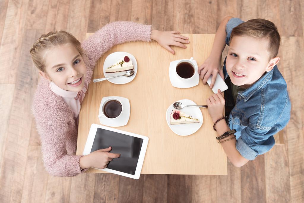 dzieci patrząc na kamery, siedząc przy stole z gadżetami na powierzchni w kawiarni - Zdjęcie, obraz