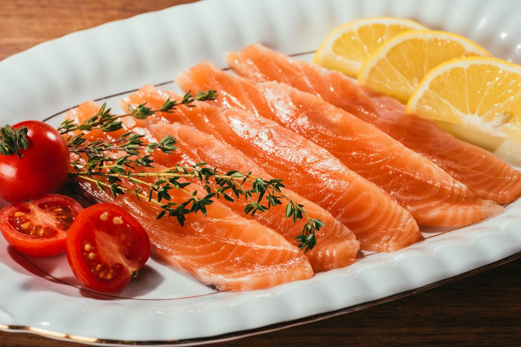 Vista ravvicinata di pezzi di pesce salmone con limone, erbe e pomodori su piatto bianco
  - Foto, immagini