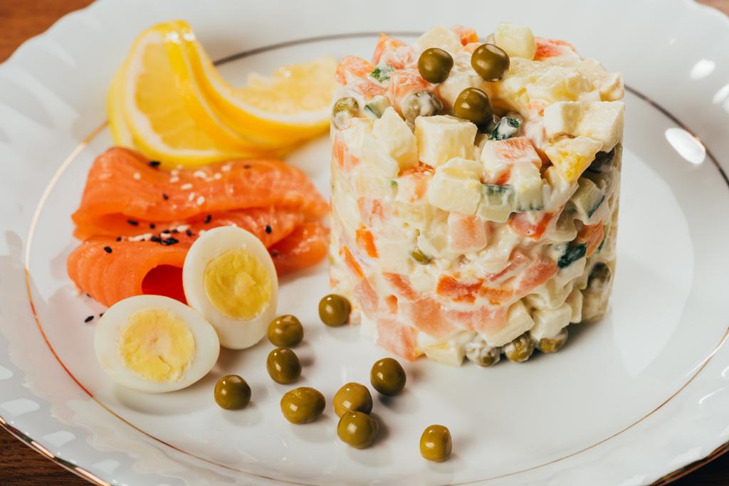 Русский салат на тарелке с разбросанным горохом, вареными яйцами и ломтиками рыбы
    - Фото, изображение