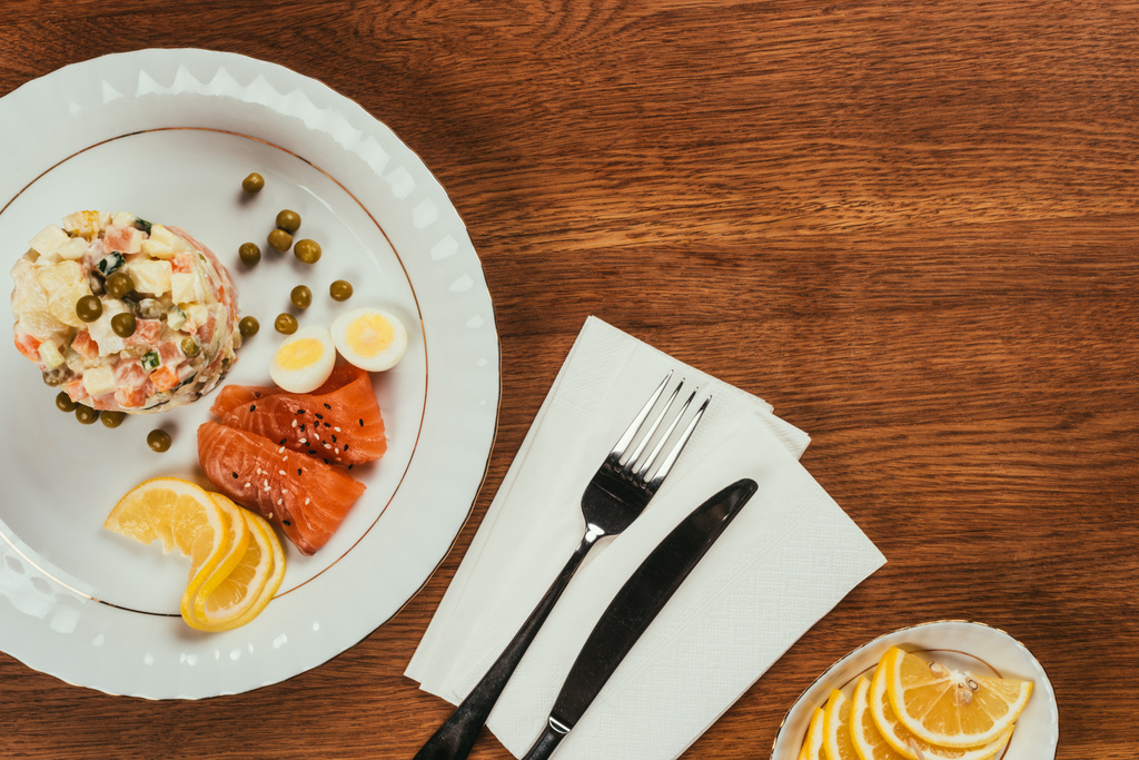 Vista superior de ensalada rusa servida con salmón y limón en plato blanco sobre mesa de madera
 - Foto, imagen