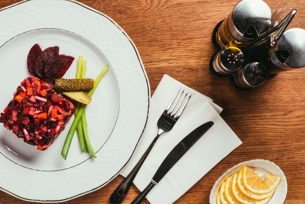 Vista superior de la ensalada Vinegret servida con cebolla verde y pepino en vinagre en plato blanco con dispensadores de condimentos en mesa de madera
 - Foto, Imagen