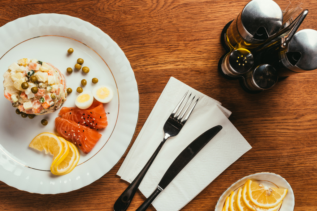 Russischer Salat auf Teller mit Erbsen, gekochten Eiern und Fischscheiben über Tisch mit Gabel und Messer auf Serviette - Foto, Bild