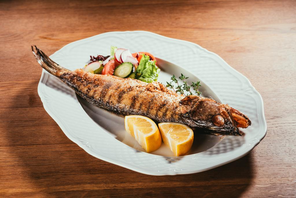 Baked fish with lemon and herbs on white plate with salad on wooden table - Photo, Image