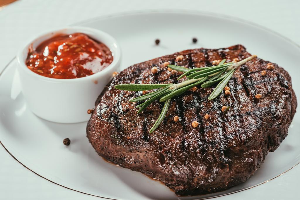 Close-up view of grilled steak with pepper, rosemary and sauce on white plate - Photo, Image