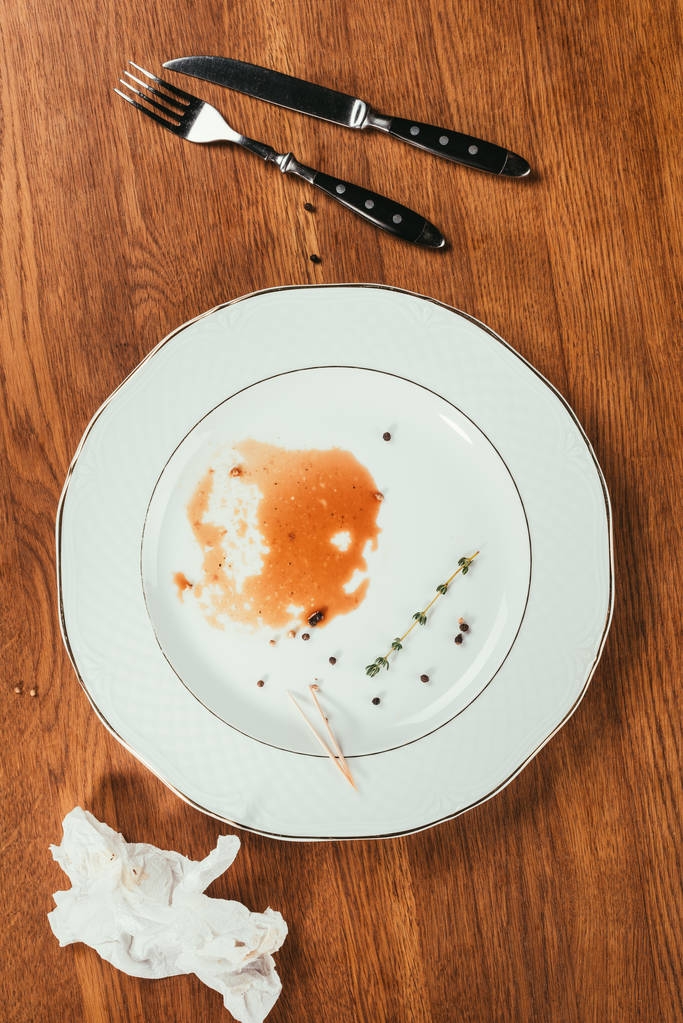 остатки мяса в почти пустой тарелке на деревянной поверхности вилкой, ножом и морщинистой салфеткой
 - Фото, изображение