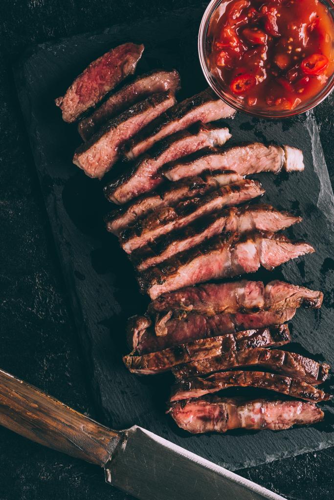 felülnézet ízletes szeletelt grillezett hús mártással és kés, fekete pala táblán   - Fotó, kép