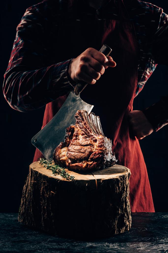 обрезанный снимок шеф-повара в фартуке с мясным ножом и вкусными ребрышками на деревянном пне
  - Фото, изображение