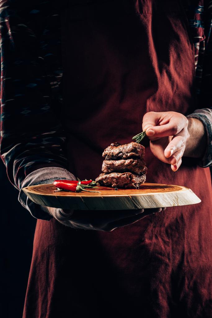 przycięte zdjęcie szefa kuchni w fartuch, trzymając deska z pyszne mięso z grilla, chili pieprz i rozmaryn  - Zdjęcie, obraz