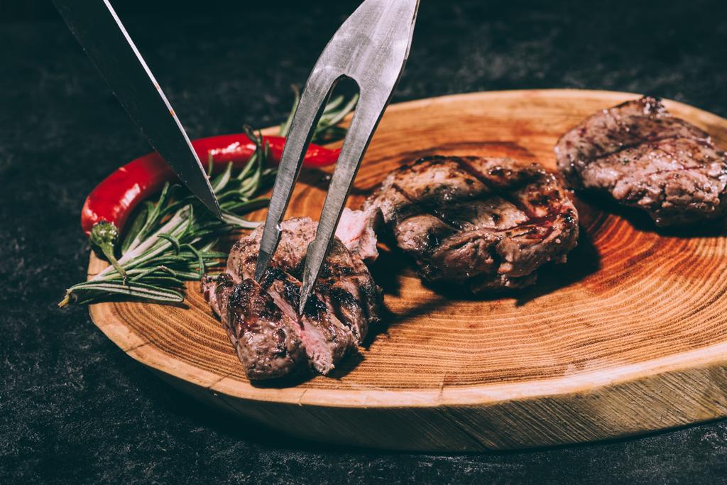 Vista ravvicinata di carne forchetta e coltello, deliziose bistecche alla griglia con rosmarino e peperoncino su tavola di legno
 - Foto, immagini