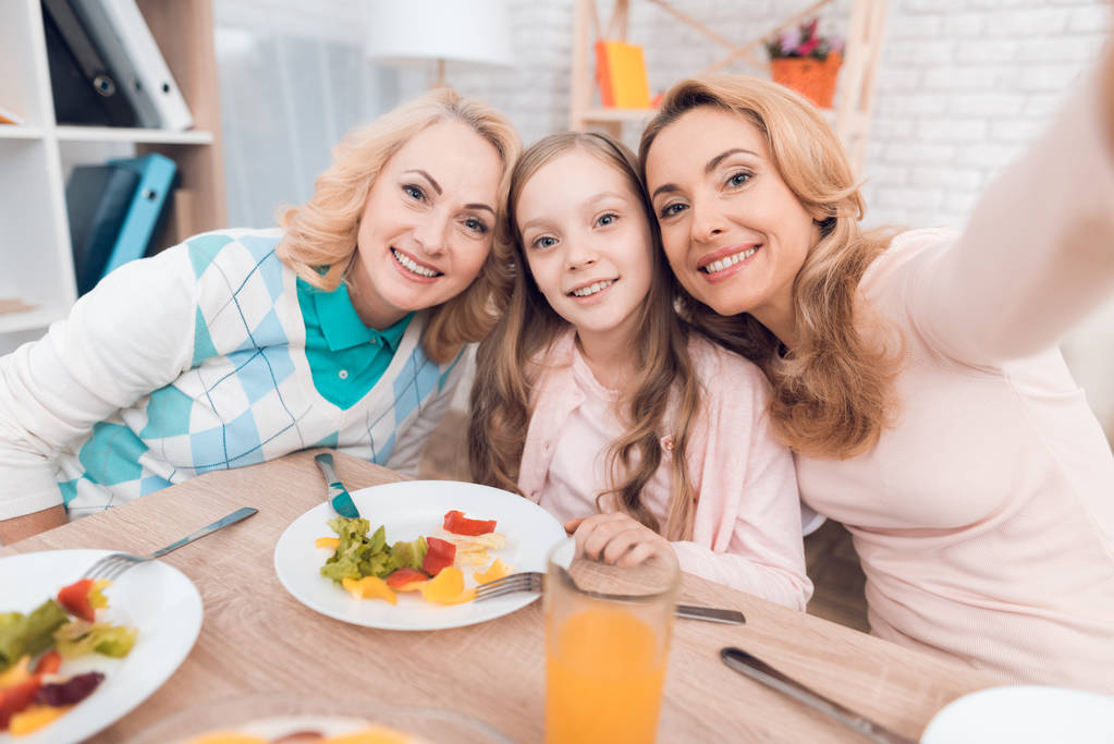 Mom, grandmother and granddaughter do selfie at the dinner table. They have dinner at home. They have vegetables and sweets on their table. - Photo, Image