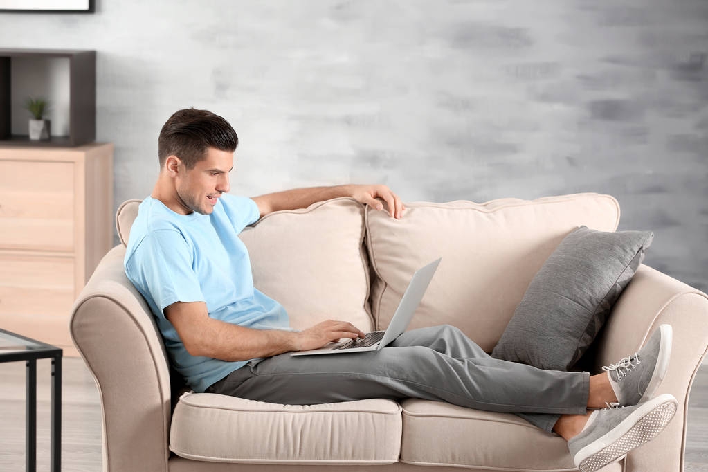 Evde koltukta istirahat ederken dizüstü bilgisayar kullanan yakışıklı adam - Fotoğraf, Görsel