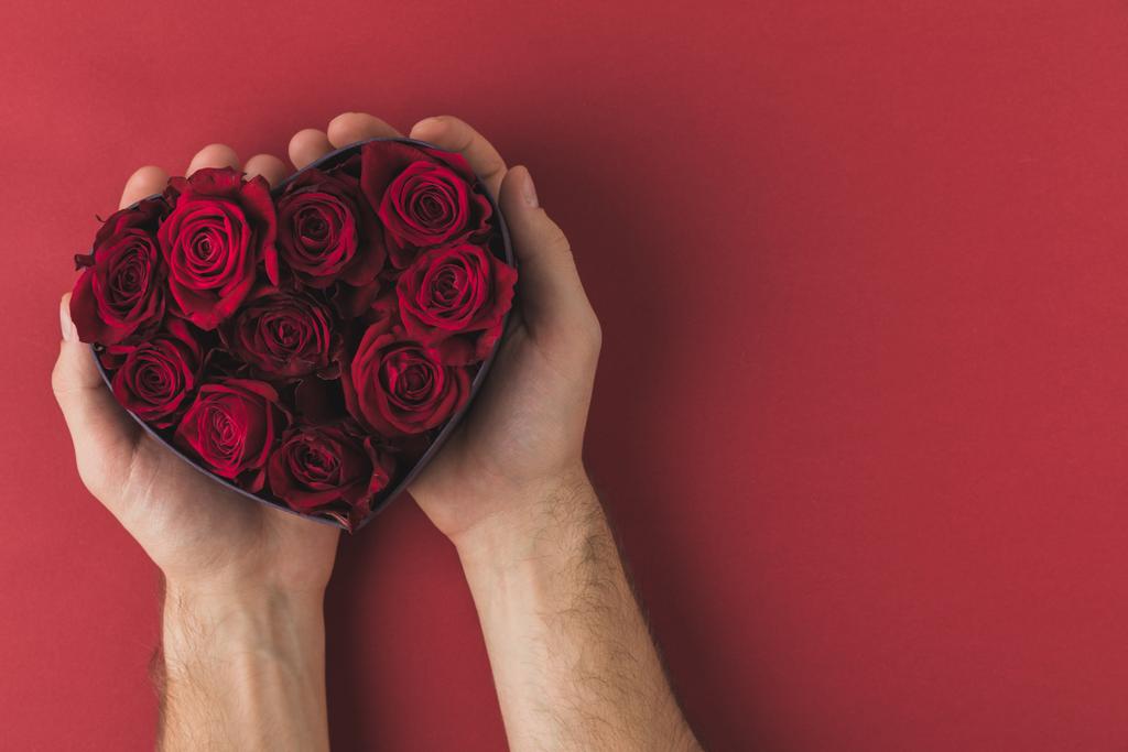 abgeschnittene Aufnahme eines Mannes mit Rosen in herzförmiger Schachtel auf roter Tischplatte, Valentinstag-Konzept - Foto, Bild