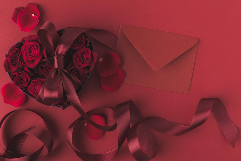 top view of roses in heart shaped gift box with ribbon and envelope isolated on red, st valentines day holiday concept - Photo, Image
