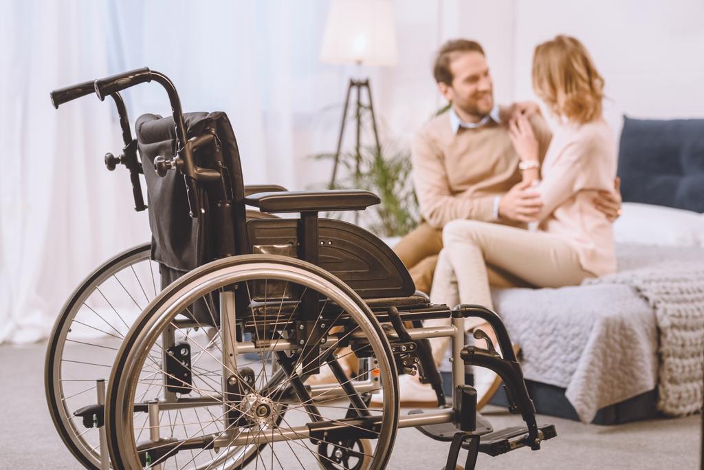 šťastný manžel s handicapem a manželka sedí a objímá na posteli s vozíkem na popředí - Fotografie, Obrázek