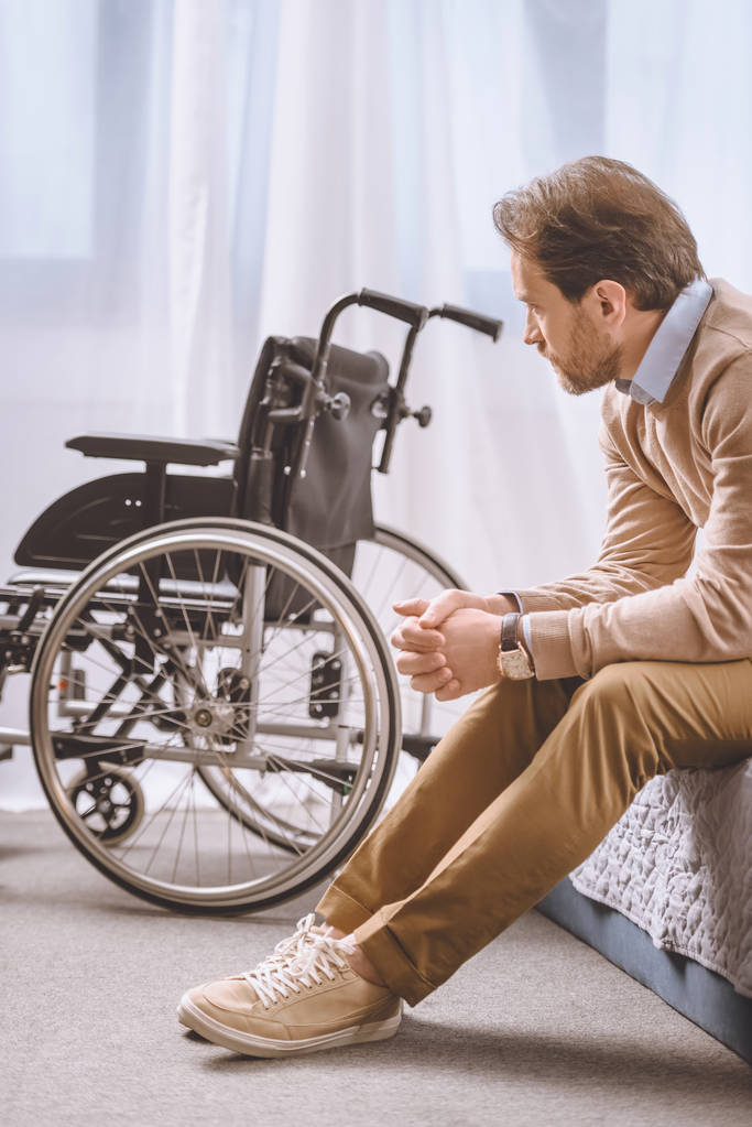 ανήσυχος άνθρωπος με αναπηρία κάθεται στο κρεβάτι και κοιτάζοντας με αναπηρικό καροτσάκι - Φωτογραφία, εικόνα