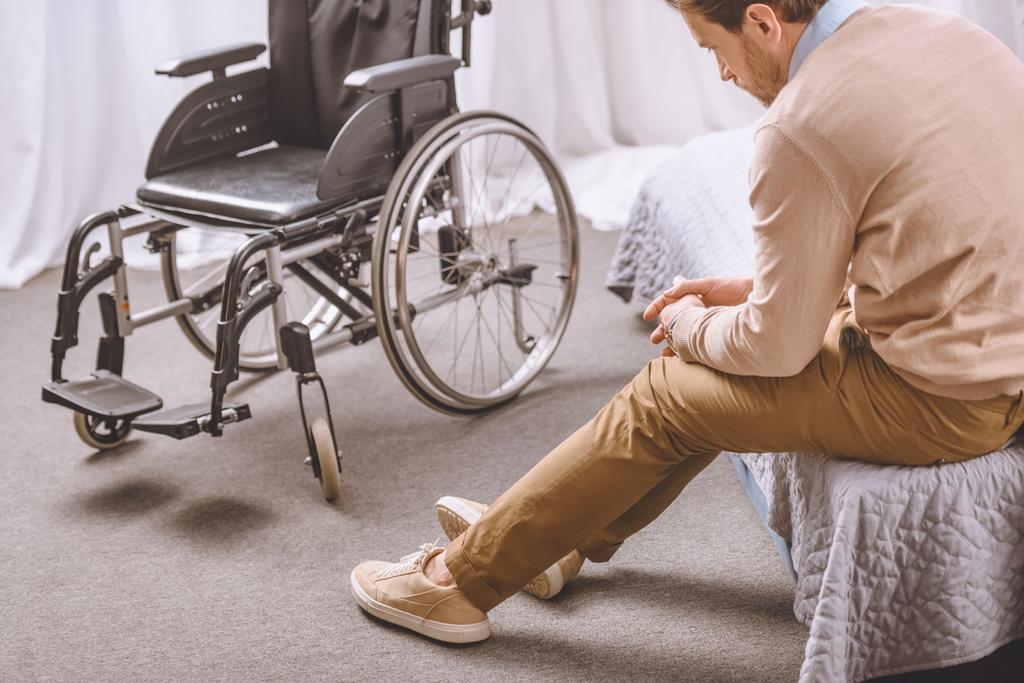 λυπημένο άνθρωπο με αναπηρία που κάθεται στο κρεβάτι, αναπηρική καρέκλα στέκεται κοντά - Φωτογραφία, εικόνα
