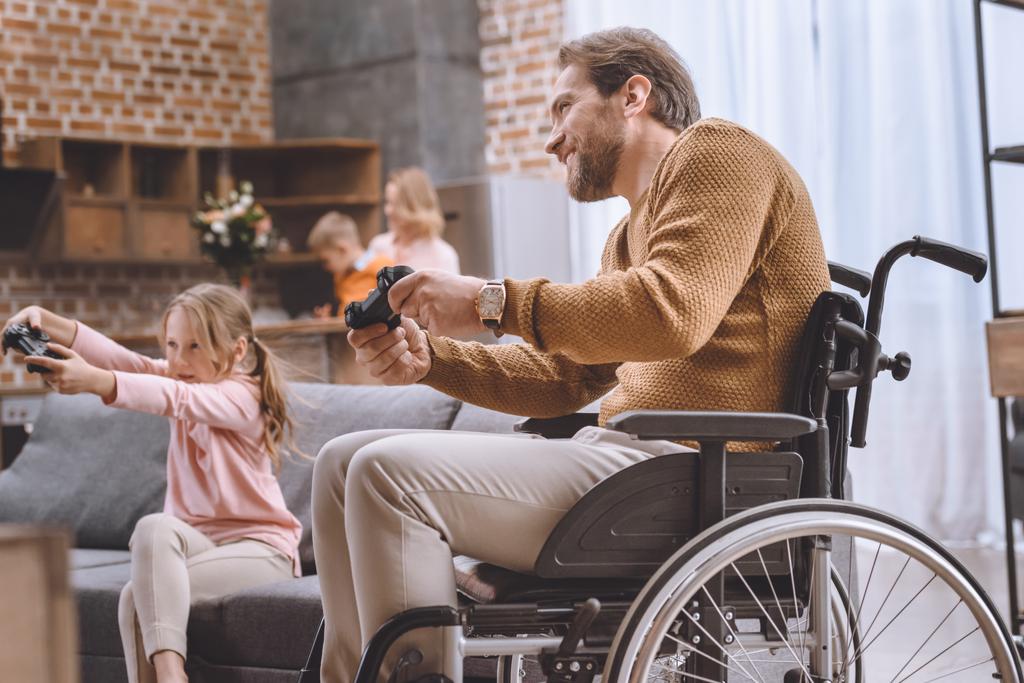 ευτυχισμένη κόρη και πατέρας σε αναπηρικό καροτσάκι να παίζει video game με χειριστήρια  - Φωτογραφία, εικόνα