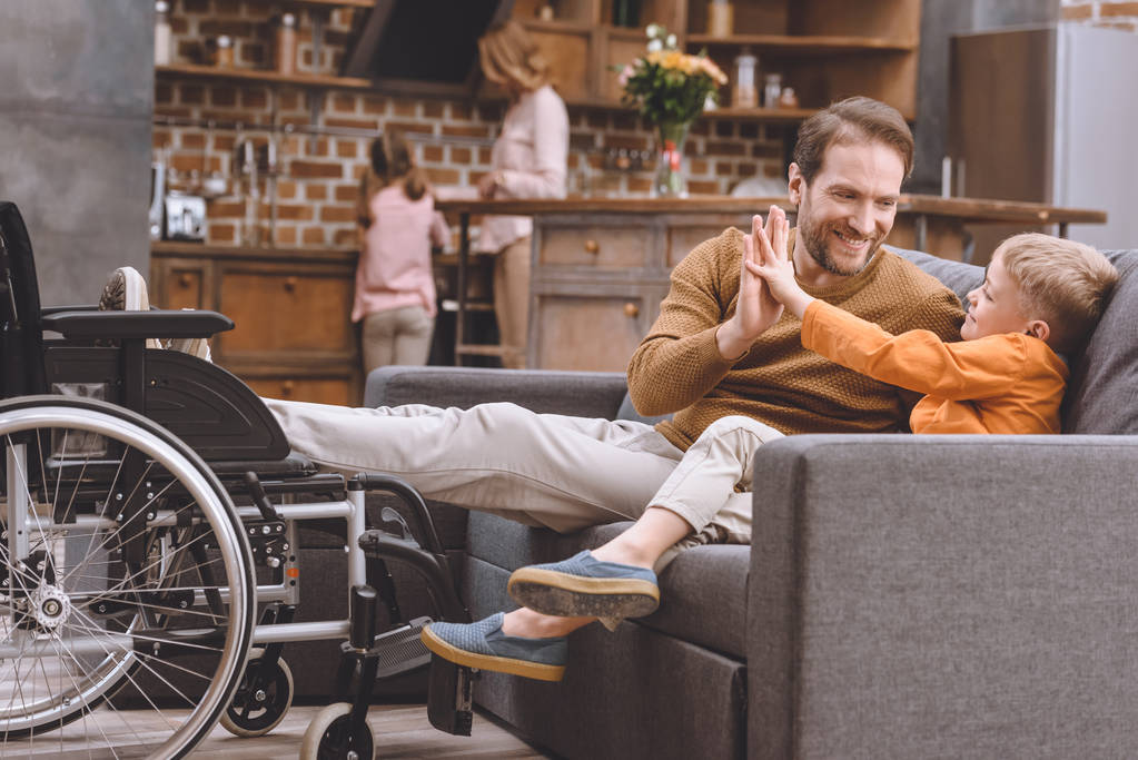 щасливий маленький син з усміхненим батьком з інвалідністю дає високі п'ять і інвалідне крісло, що стоїть біля дивана
 - Фото, зображення
