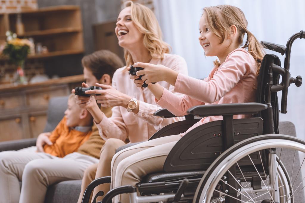 glückliche Familie mit behindertem Kind im Rollstuhl, das zu Hause mit Joysticks spielt  - Foto, Bild