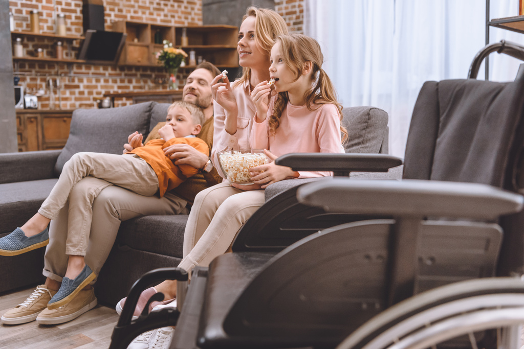 lähikuva pyörätuolista ja perheestä istuu sohvalla ja syö popcornia takana
 - Valokuva, kuva