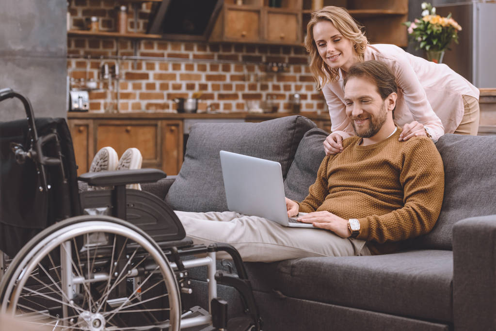 ευτυχισμένη γυναίκα που βλέπουν άτομα με ειδικές ανάγκες σύζυγος, βάζοντας τα πόδια σε αναπηρικό καροτσάκι και να χρησιμοποιούν φορητό υπολογιστή στο σπίτι - Φωτογραφία, εικόνα