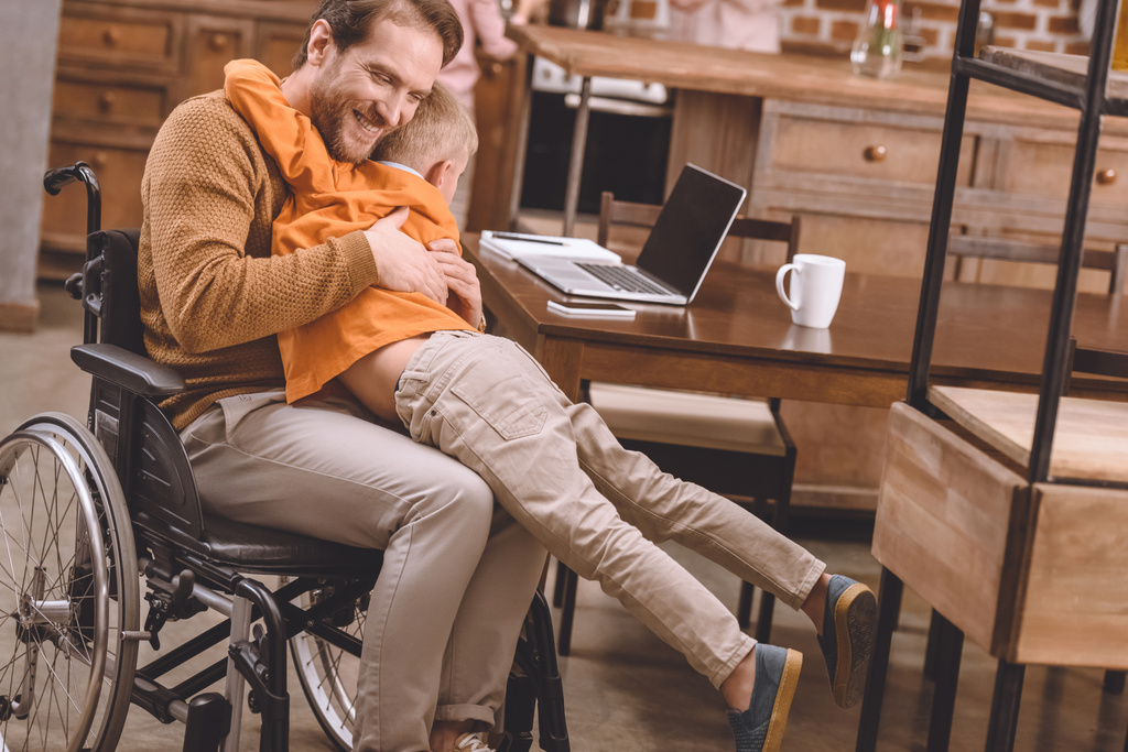счастливый отец-инвалид в инвалидной коляске обнимается с милым маленьким сыном дома
 - Фото, изображение