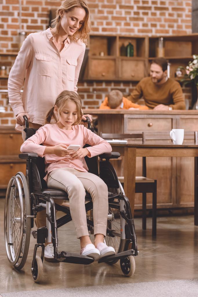 Χαμογελώντας η μητέρα βλέπει ανάπηρη κόρη κάθεται σε αναπηρικό καροτσάκι και χρησιμοποιώντας το smartphone στο σπίτι   - Φωτογραφία, εικόνα