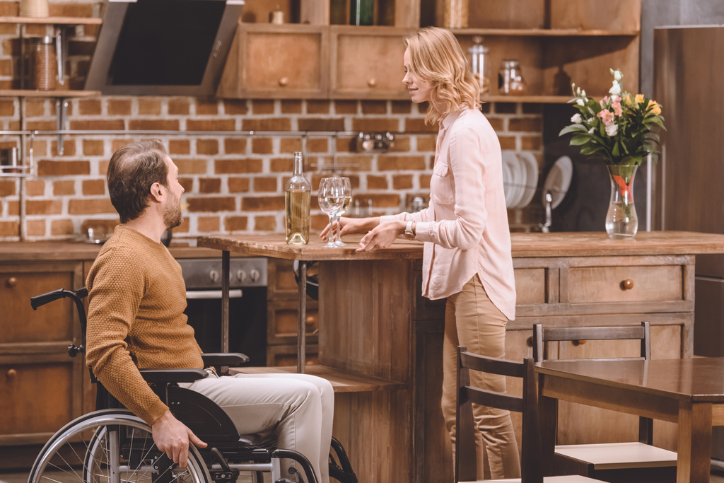 Seitenansicht einer Frau mit Weinflasche und Gläsern, die zu Hause einen behinderten Mann im Rollstuhl ansieht - Foto, Bild