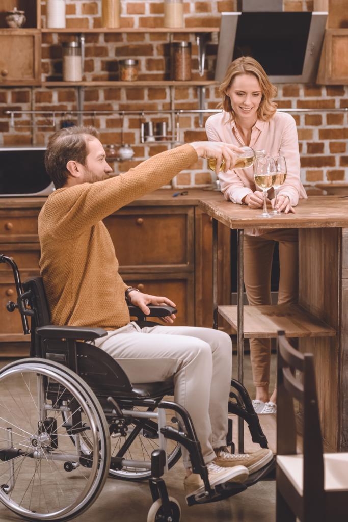muž v invalidním vozíku nalévání vína a usmívající se žena hospodářství brýle doma - Fotografie, Obrázek