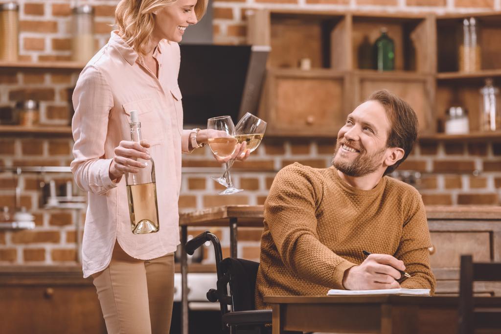 mosolygó nő bor gazdaság szemüveg és üveg, míg a fogyatékkal élő férje tolószékben jegyzeteket otthon   - Fotó, kép