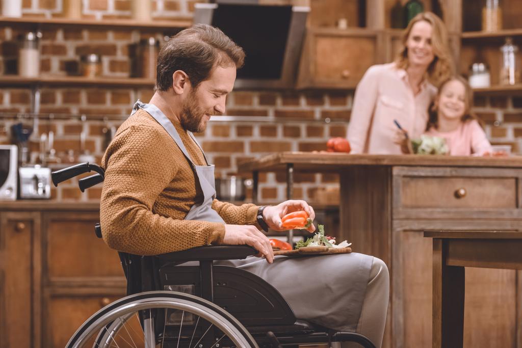 vista lateral del hombre sonriente en silla de ruedas cortando verduras para ensalada, mientras que la madre y la hija feliz de pie detrás
 - Foto, imagen