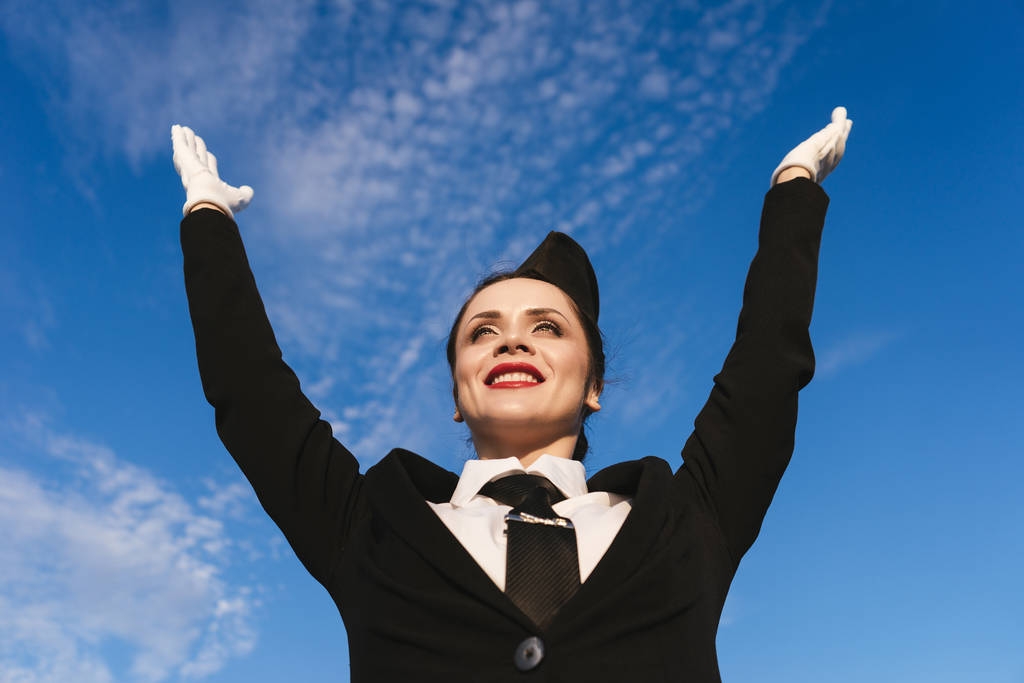 feliz mujer sonriente azafata en uniforme levantó las manos, contra el cielo azul, esperando el avión
 - Foto, imagen