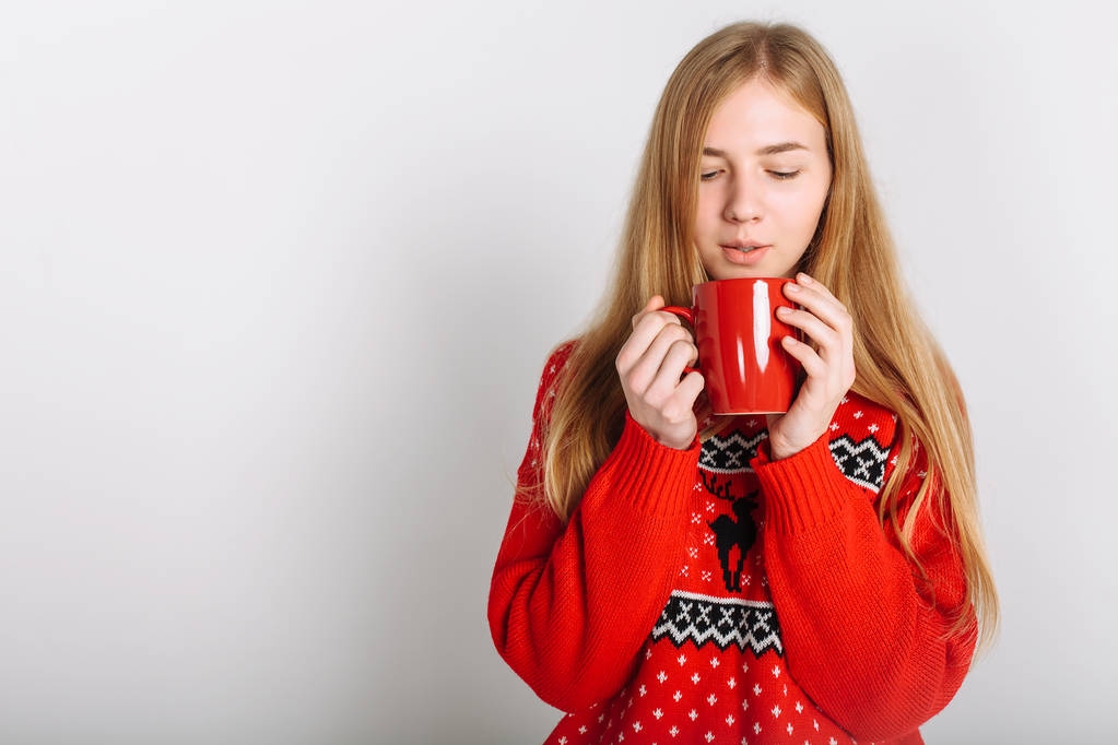 Κορίτσι με ένα πουλόβερ Χριστουγέννων ή Πρωτοχρονιάς στο Studio κρατώντας ένα φλιτζάνι αρωματικό καφέ. Απομονωμένο μέρος για διάθεση των Χριστουγέννων κείμενο. - Φωτογραφία, εικόνα