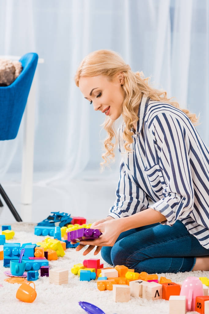 Vista lateral da jovem mãe com brinquedos sentados no chão em casa
 - Foto, Imagem