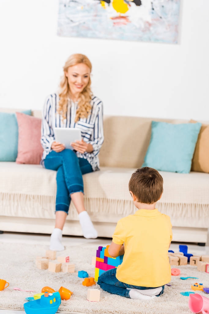 маленький мальчик играет с игрушками в то время как мать использует планшет на диване дома
 - Фото, изображение