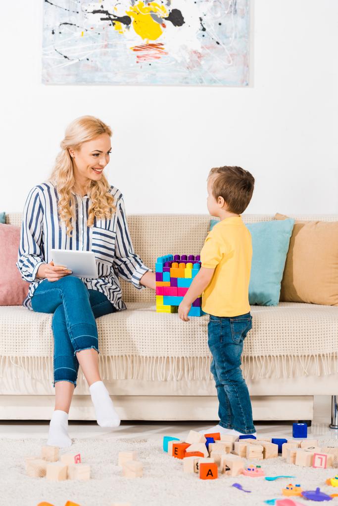 little boy showing toys to smiling mother with tablet on sofa at home - Photo, Image