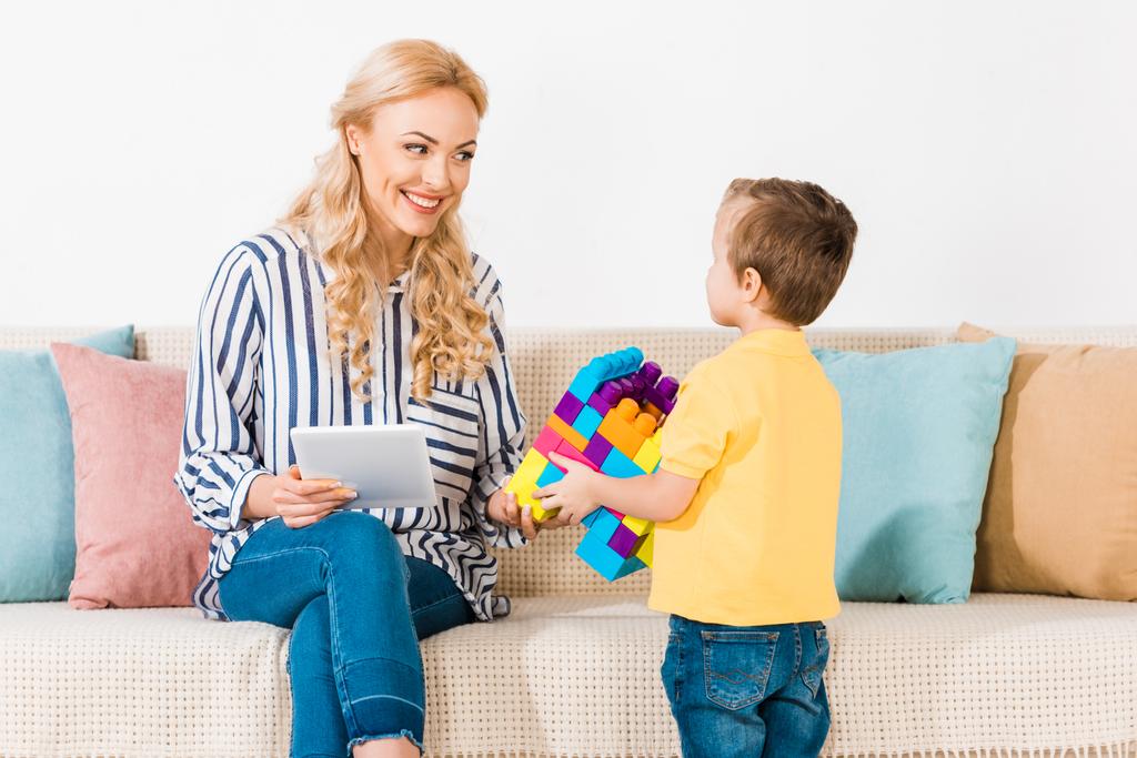 niño pequeño mostrando juguetes a la madre sonriente con la tableta en el sofá en casa
 - Foto, Imagen