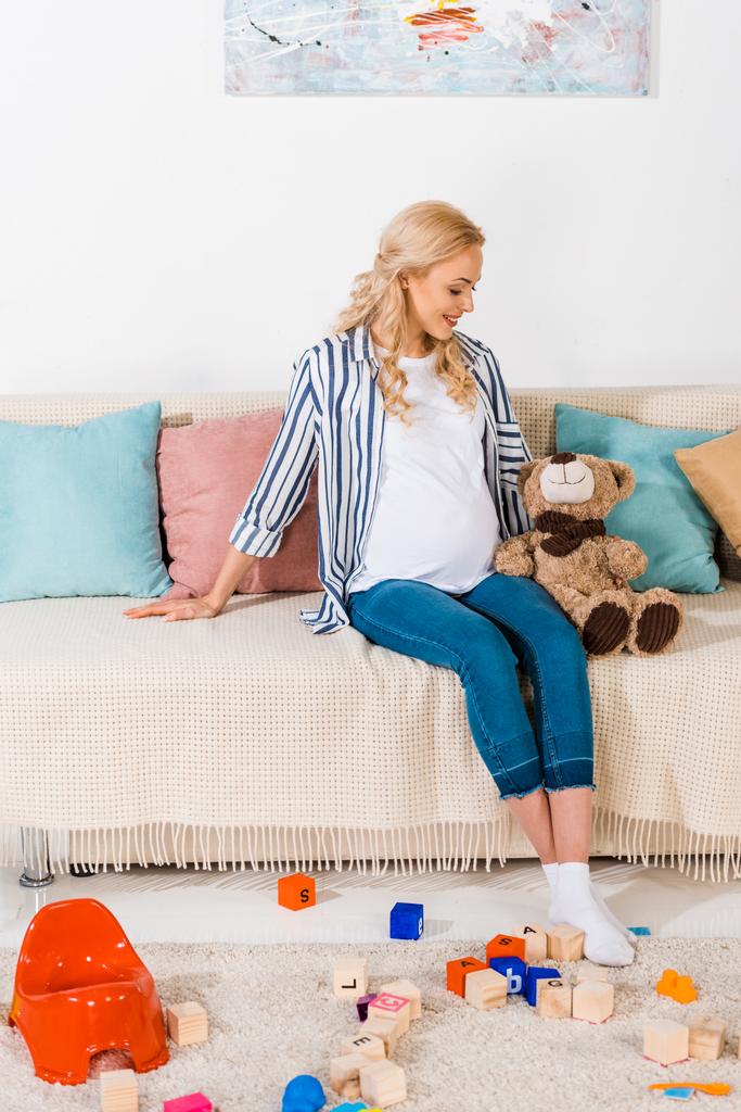 Lächelnde Schwangere sitzt mit Teddybär auf Sofa - Foto, Bild