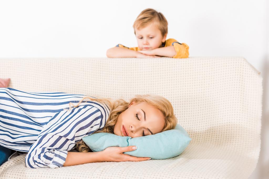 cute little boy looking at mother sleeping on sofa at home - Photo, Image