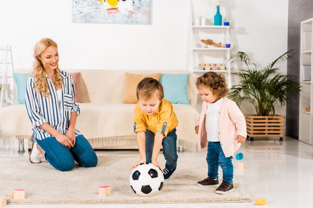 Улыбающаяся мать смотрит на милых маленьких детей, играющих в футбол дома
 - Фото, изображение