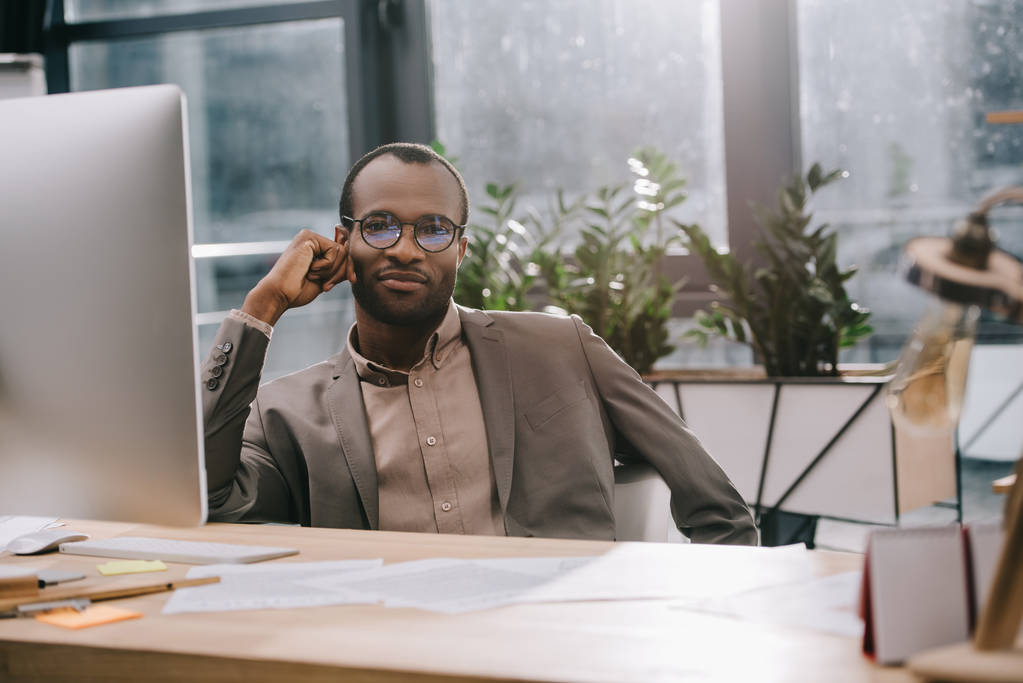 pěkný africký americký podnikatel sedí na pracovišti a při pohledu na fotoaparát - Fotografie, Obrázek
