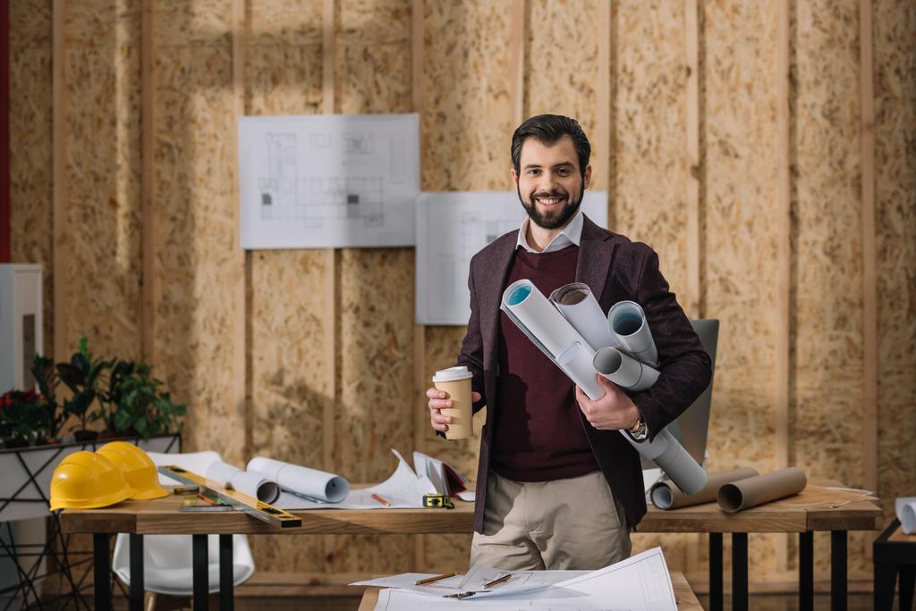 schöner junger Architekt mit Coffee to go und gerollten Entwürfen am Arbeitsplatz - Foto, Bild