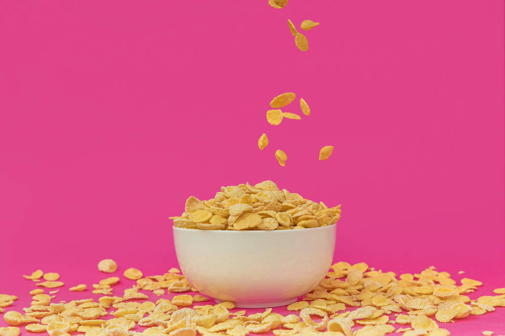 Nahaufnahme von süßen gesunden Cornflakes, die in eine weiße Schüssel auf rosa fallen  - Foto, Bild