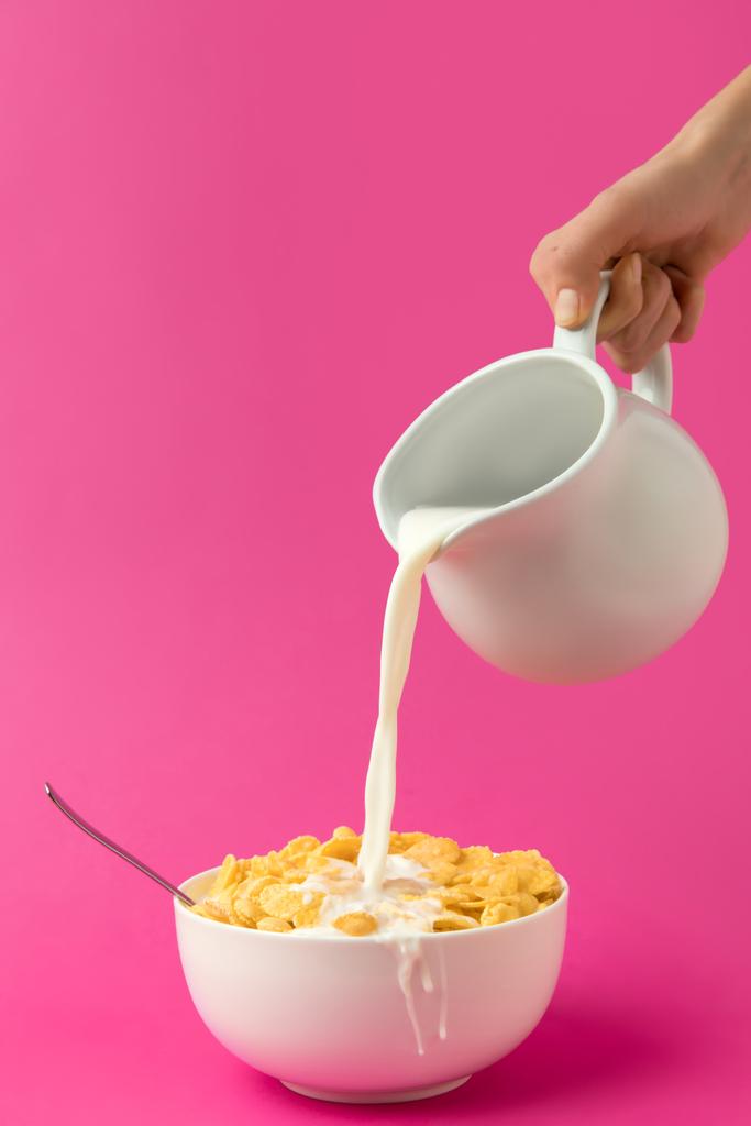 cropped shot of hand holding jug and pouring milk into bowl with corn flakes isolated on pink - Photo, Image