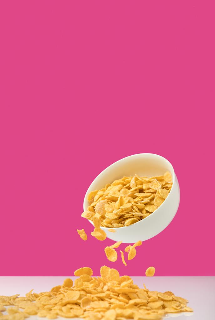 Nahaufnahme einer weißen Schüssel mit Cornflakes, die sich auf rosa ausschütten  - Foto, Bild