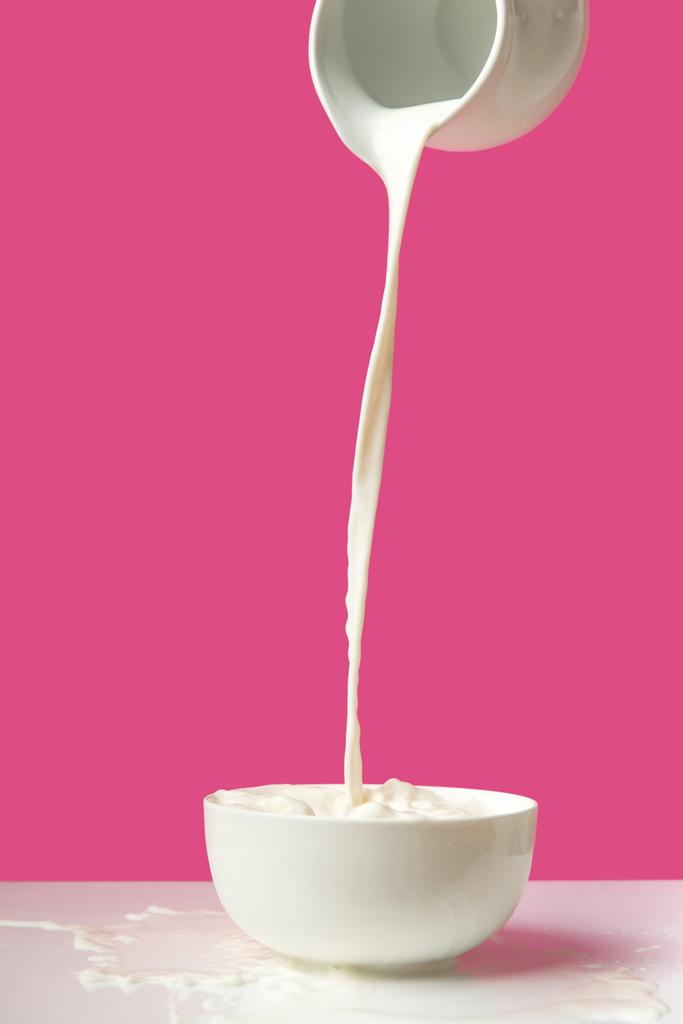 Frische gesunde Milch aus Krug in Schüssel auf rosa gießen     - Foto, Bild