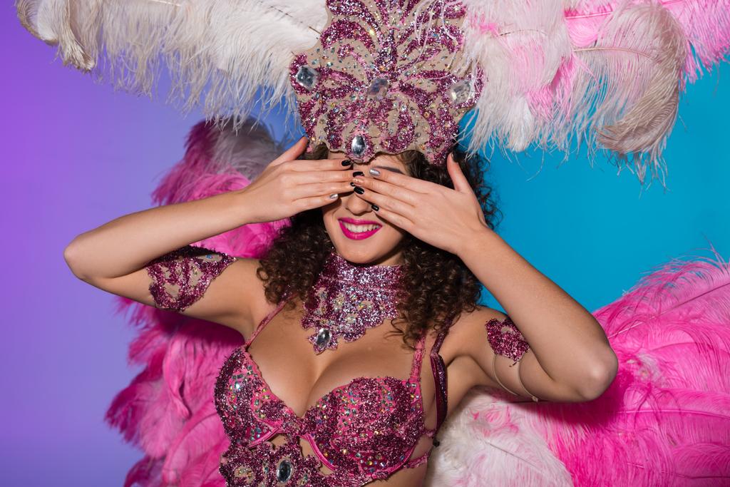 Mujer alegre disfrazada de carnaval con plumas rosas cubriendo sus ojos aislados sobre fondo azul
 - Foto, imagen