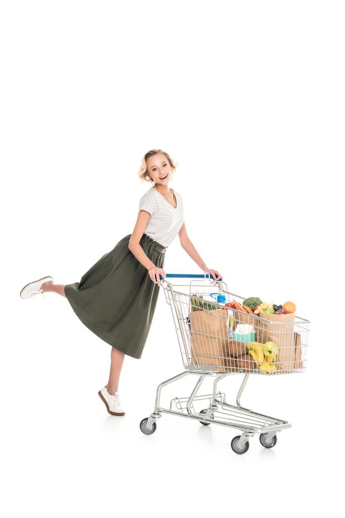 Szczęśliwa młoda kobieta stojąc z wózka na zakupy i uśmiecha się do kamery na białym tle     - Zdjęcie, obraz