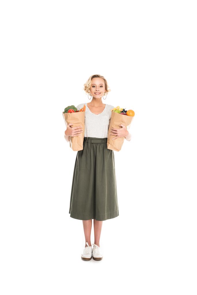 full length view of woman holding paper bags with fruits and vegetables and smiling at camera isolated on white - Photo, Image